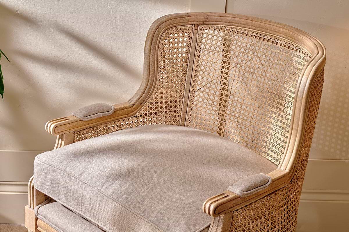 The Kaziria Cane & Linen Armchair by nkuku features natural linen, a light mango wood frame, and woven cane back and sides. Its beige cushion and padded armrests offer comfort, perfect for any space with a wooden floor and cream wall.