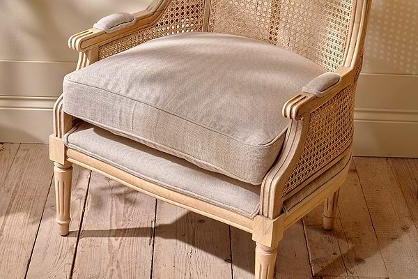 The Kaziria Cane & Linen Armchair - Natural by nkuku features a beige cushion and woven cane back and sides. Crafted from mango wood, it sits on light floorboards against a cream wall with sunlight casting patterned shadows.