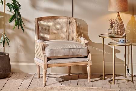 The Kaziria Cane & Linen Armchair by nkuku, featuring a woven back, sides, beige cushion, and armrest pads, sits on a wooden floor beside a round gold side table with a lamp, dried flowers in a vase, and a cup. Sunlight brightens the room and nearby potted plant.