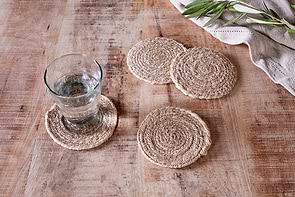 A glass of water sits on an nkuku Keso Woven Coaster—Natural (Set of 4)—atop a rustic wooden table. Three empty coasters are nearby, with a folded beige cloth and greenery sprigs in the upper right corner.