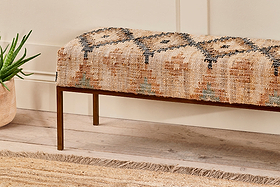 The Kesu Kilim & Iron Bench by nkuku features vintage-style woven fabric in warm beige, rust, and black on slim bronze metal legs. Shown on light wood flooring with a jute rug and a potted aloe plant.