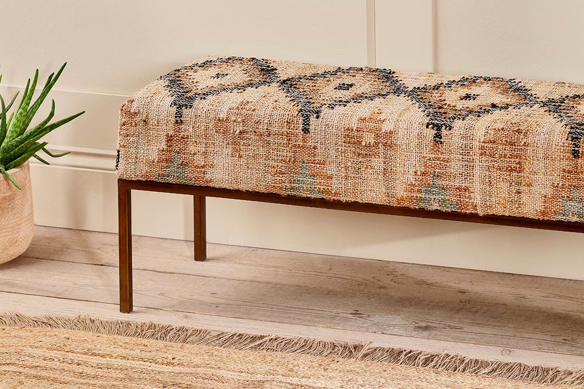 The Kesu Kilim & Iron Bench by nkuku features vintage-style woven fabric in warm beige, rust, and black on slim bronze metal legs. Shown on light wood flooring with a jute rug and a potted aloe plant.