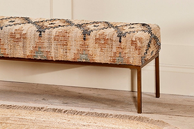 The Kesu Kilim & Iron Bench by nkuku features a cushioned seat in handloom woven fabric with beige, brown, black, and light blue patterns. It stands on slim iron legs beside a textured beige rug and pale wall on a light wooden floor.