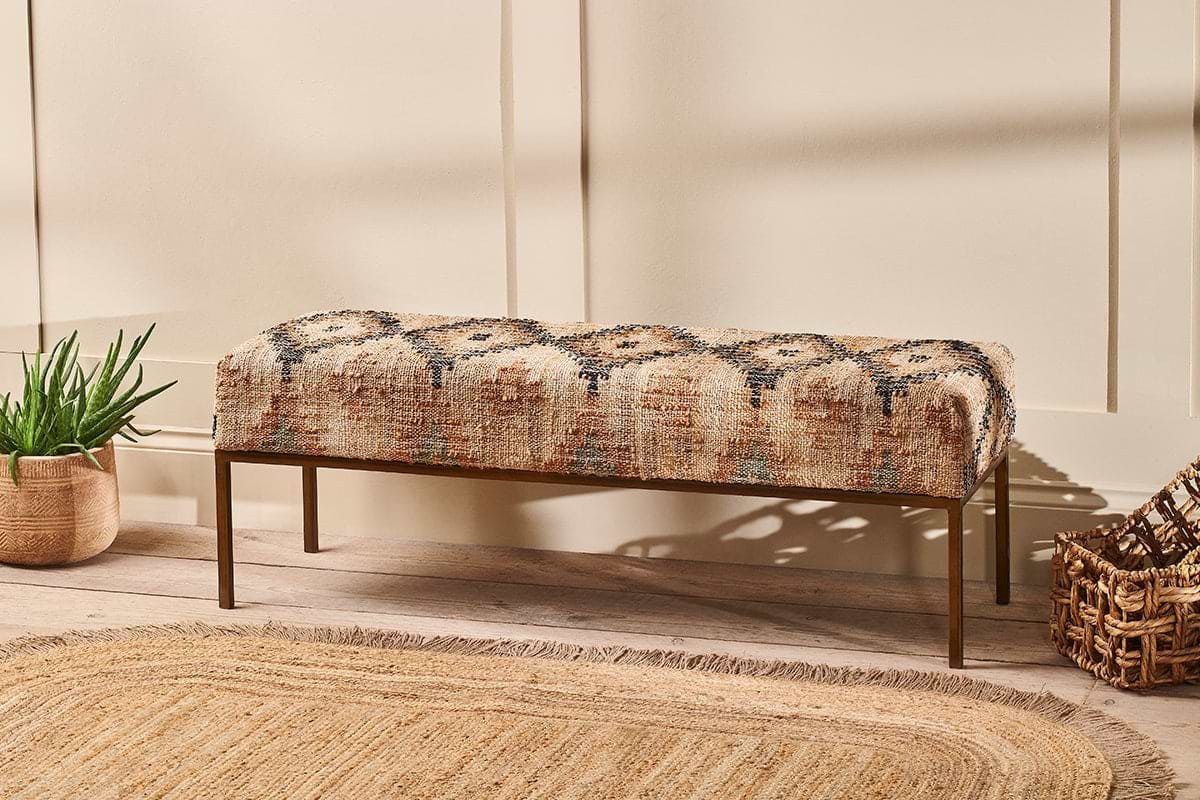 The nkuku Kesu Kilim & Iron Bench, featuring geometric beige, brown, and blue fabric, sits against a cream wall beside a potted plant and woven basket. A round jute rug on the light wood floor completes this warm, inviting scene.