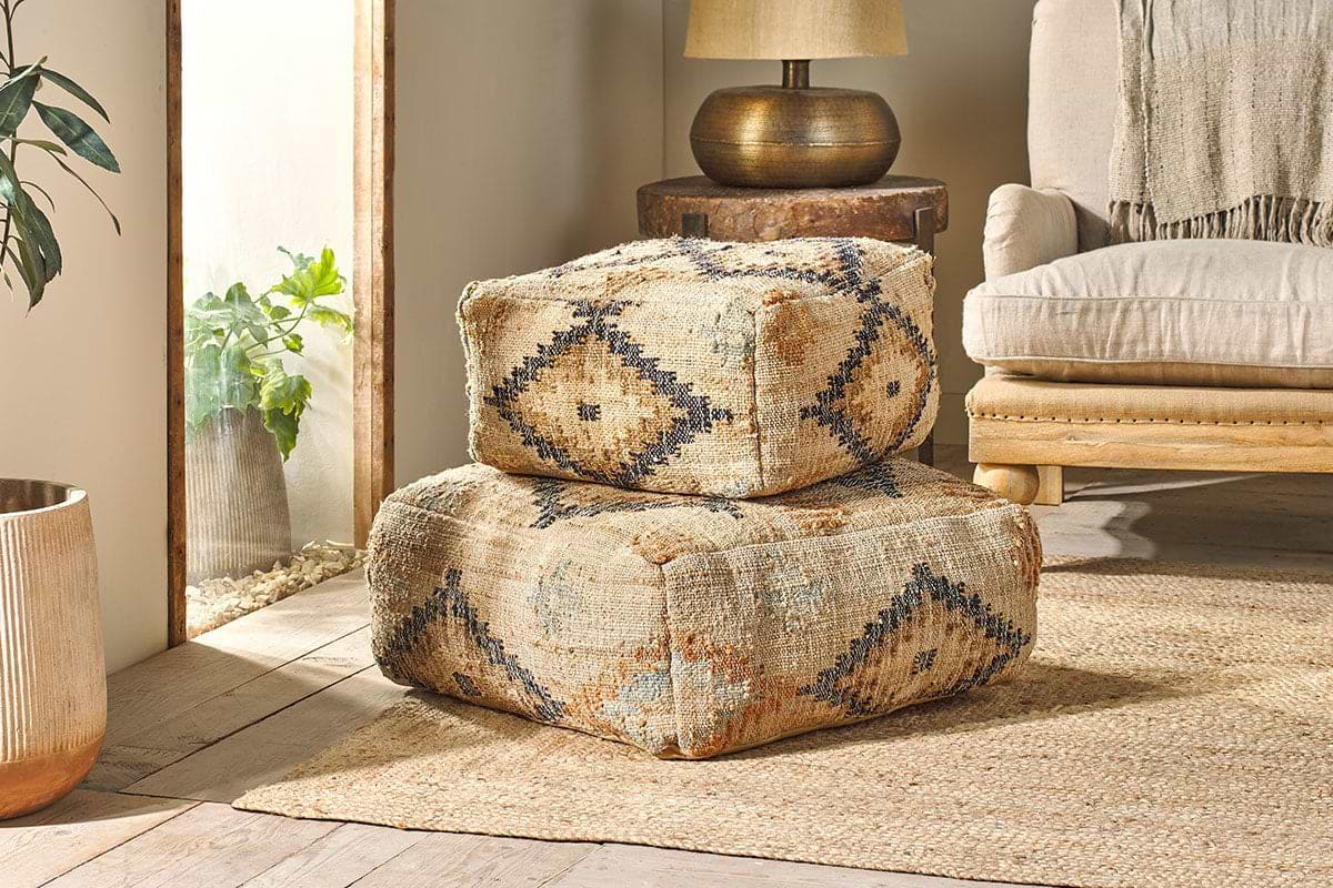 Two large Kesu Kilim Poufs by nkuku, featuring geometric diamond patterns in earthy tones, are stacked in a bright living room with a woven rug, beige armchair, wooden side table with lamp, and potted plant by a window.