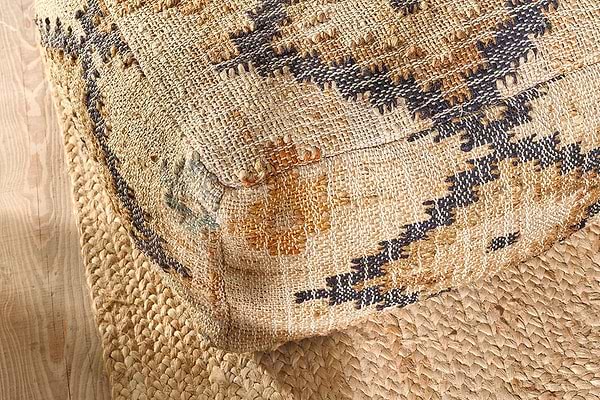 The nkuku Kesu Kilim Pouf - Large, featuring tan, black, and mustard geometric patterns, sits on a natural braided jute rug over light wood floors, accentuating a rustic bohemian vibe with its woven texture and bold design.