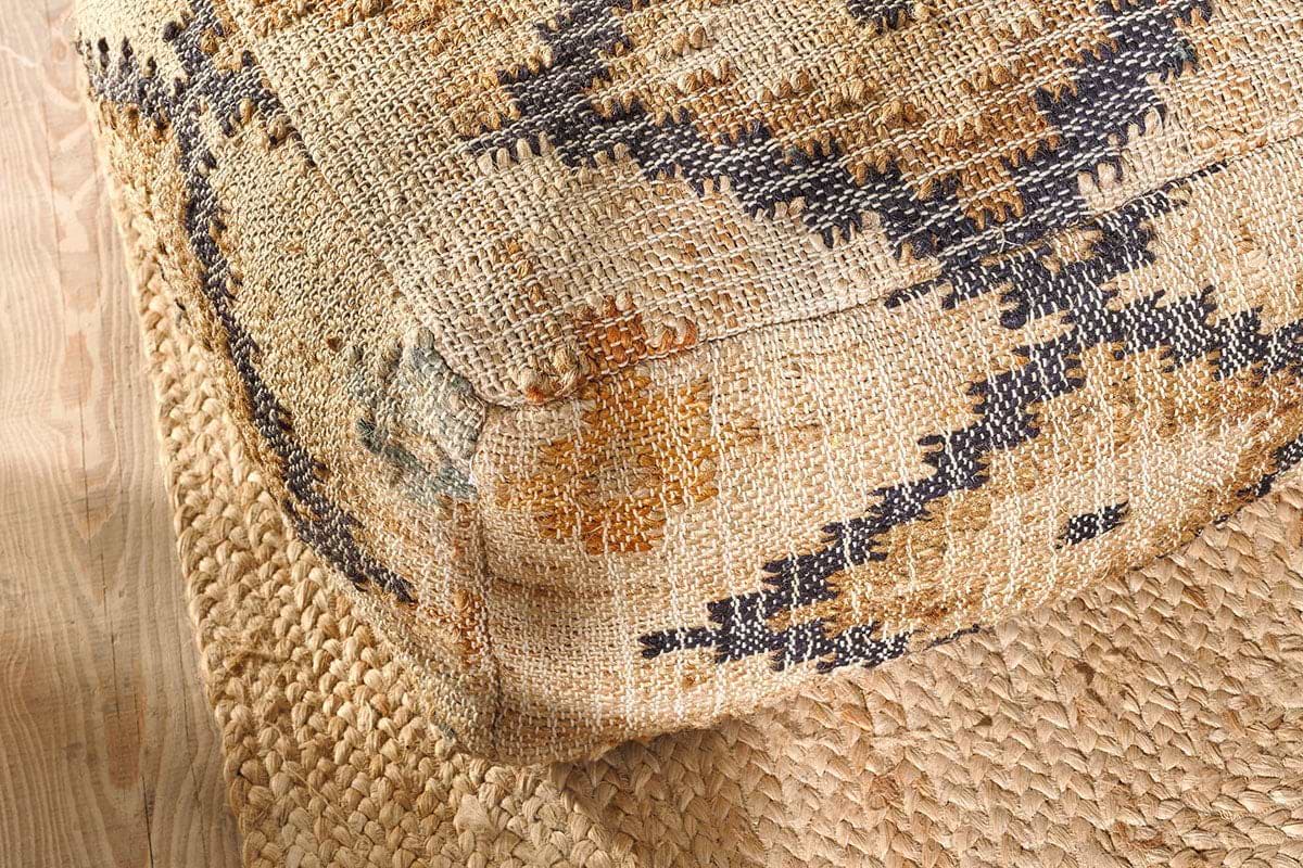The nkuku Kesu Kilim Pouf - Large, featuring tan, black, and mustard geometric patterns, sits on a natural braided jute rug over light wood floors, accentuating a rustic bohemian vibe with its woven texture and bold design.