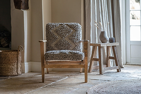 A nkuku Keya Chunky Weave & Mango Wood Chair - Natural, with a thick gray cushion, sits by the window on a stone floor. A small wooden table with a ribbed vase and dried plants completes the cozy, rustic look.