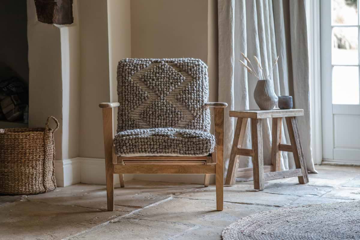 A nkuku Keya Chunky Weave & Mango Wood Chair - Natural, with a thick gray cushion, sits by the window on a stone floor. A small wooden table with a ribbed vase and dried plants completes the cozy, rustic look.