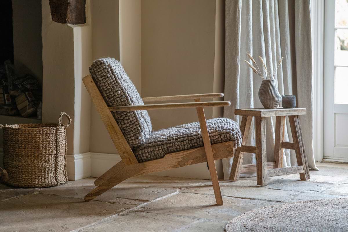A cozy room with the nkuku Keya Chunky Weave & Mango Wood Chair in natural, gray wool cushions, a woven basket, and rustic wood side table with dried plants. Beige curtains filter sunlight onto stone tile floors and a round rug.