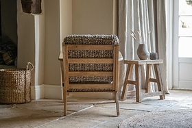 A nkuku Keya Chunky Weave & Mango Wood Chair - Natural sits in a cozy room beside a small wooden table with a ceramic vase of dried grass; a woven basket rests on the stone floor near sunlit, neutral-curtained windows.