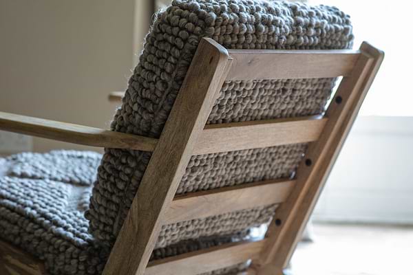 A close-up of the Keya Chunky Weave & Mango Wood Chair - Natural by nkuku shows textured gray wool cushions and natural mango wood, softly lit by sunlight with a bright window blurred in the background.
