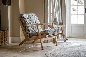 The nkuku Keya Chunky Weave & Mango Wood Chair - Natural, featuring textured grey wool upholstery and light mango wood, sits on a stone floor next to a small wooden stool topped with a vase of dried grass.