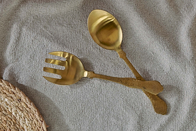 The Khana Salad Servers - Brushed Gold (Set of 2) by nkuku—a spoon and fork—rest crossed on textured gray fabric, their hammered handles radiant, with a woven placemat in the corner for an elegant table setting.
