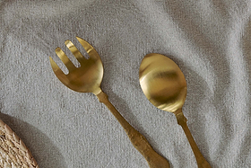 The nkuku Khana Salad Servers - Brushed Gold (Set of 2), featuring hammered handles, rest on a textured beige cloth, with a woven placemat visible in the bottom left corner.