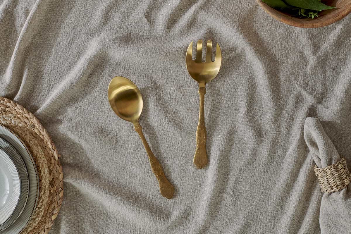 The Khana Salad Servers - Brushed Gold by nkuku, a set of two with hammered handles, rest on a beige tablecloth beside neatly stacked tableware and a napkin in a natural ring.