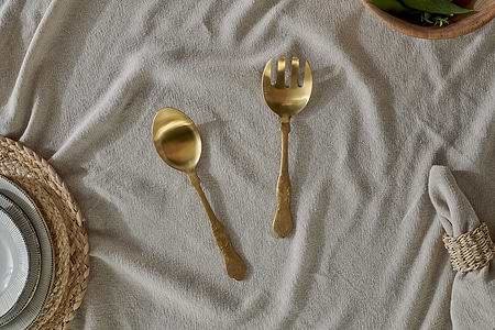 The Khana Salad Servers - Brushed Gold by nkuku, a set of two with hammered handles, rest on a beige tablecloth beside neatly stacked tableware and a napkin in a natural ring.