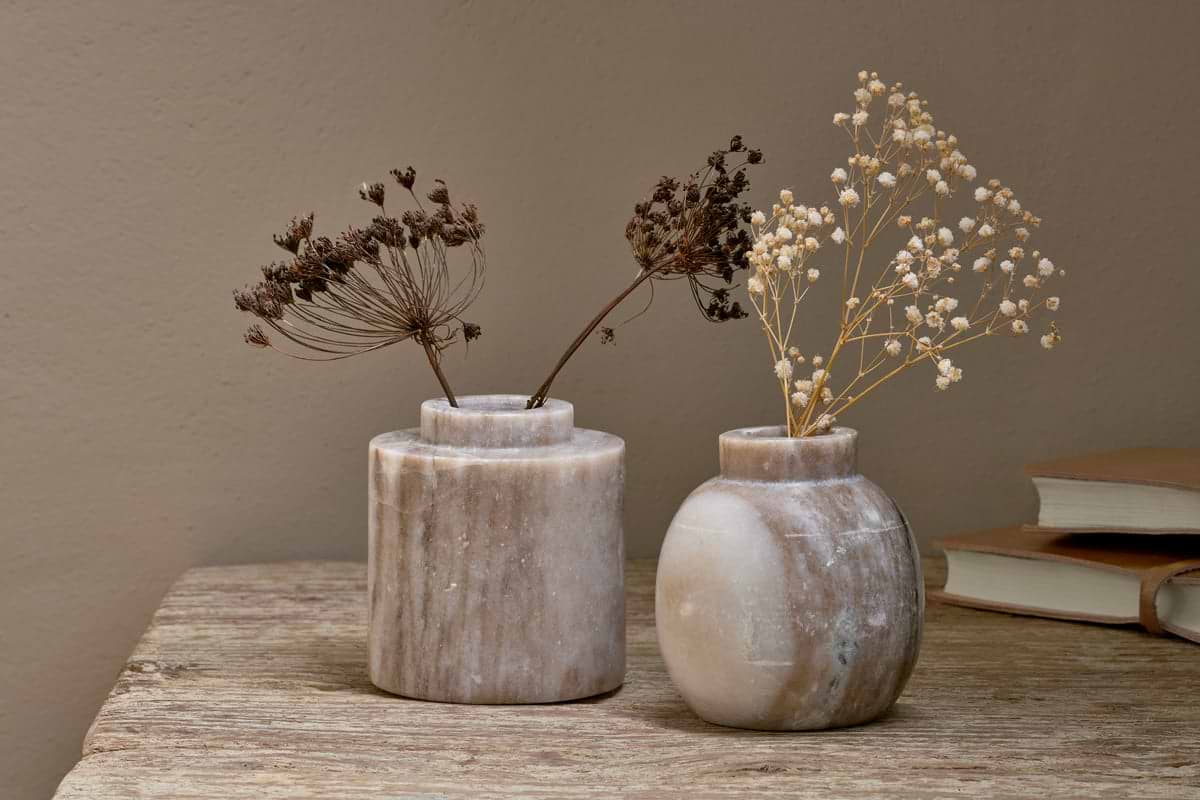 Two nkuku Khari Marble Vases in grey, each hand-carved, sit on a rustic wooden surface. One contains dried flowers with dark stems and the other with pale, delicate stems. Two closed books are visible in the background.
