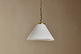 The Kiara Organic Shape Ceramic Pendant Light by nkuku hangs from the ceiling by a chain, featuring an off-white conical shade with smooth finish and antique brass trim—ideal for kitchen lighting against a neutral beige wall.