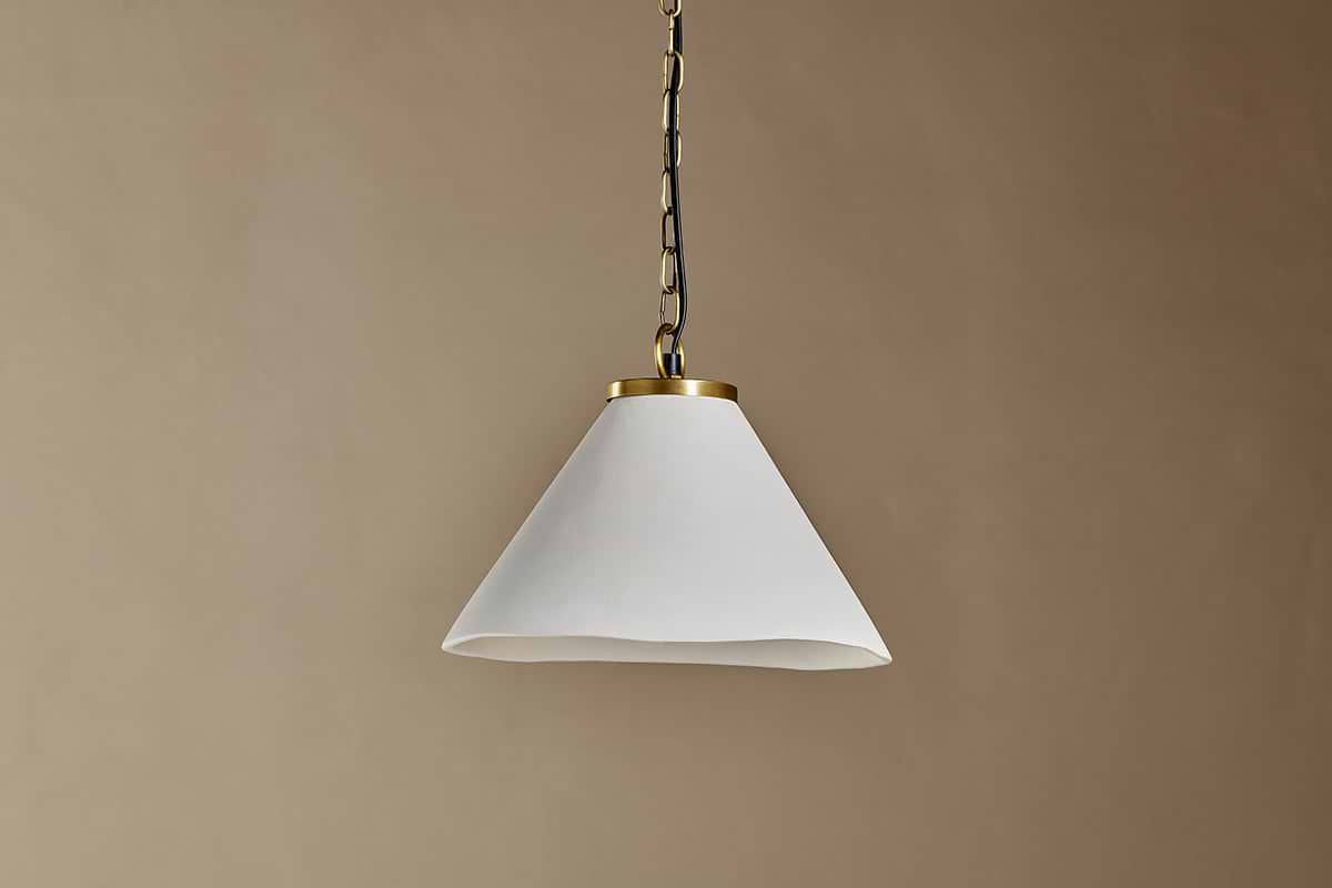 The Kiara Organic Shape Ceramic Pendant Light by nkuku hangs from the ceiling by a chain, featuring an off-white conical shade with smooth finish and antique brass trim—ideal for kitchen lighting against a neutral beige wall.