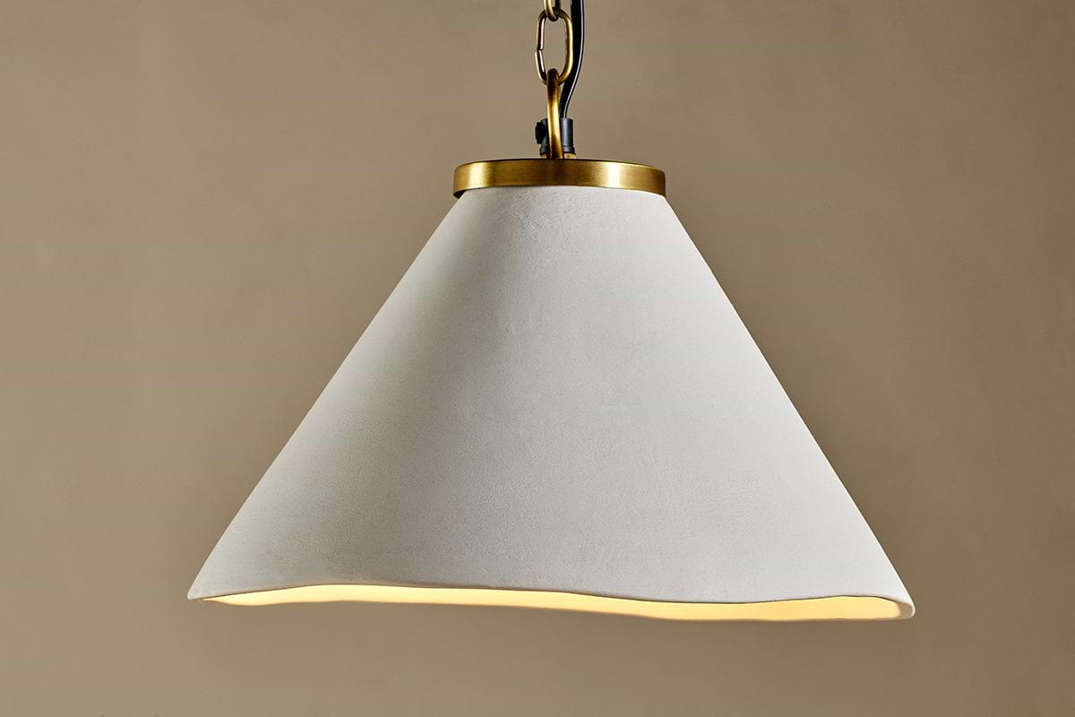The Kiara Organic Shape Ceramic Pendant Light by nkuku features a wide, wavy-edged off-white shade and antique brass chain, creating a handcrafted modern look against a taupe backdrop.