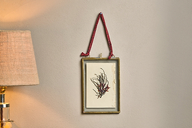 A small Kiko Brass Frame by nkuku, featuring abstract black and red strokes, hangs on a beige wall with a recycled sari tie. To the left, a lamp casts warm light, highlighting the frame’s gold finish.