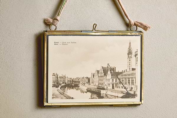 A vintage black-and-white canal scene is displayed in the Kiko Brass Frame by nkuku, which hangs on a beige wall with a pink recycled sari tie. The upper left of the photo reads: “Gent. Quai aux Herbes. Gand.—Graslei.”.