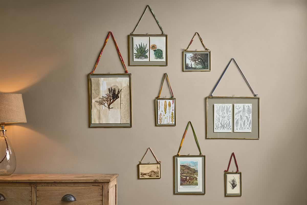 A beige wall features nine assorted-size nkuku Kiko Brass Frames, each hung with colorful recycled sari ties, displaying botanical art. A wooden dresser and a glass-based lamp are visible in the lower left corner.
