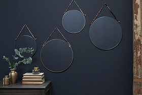 Four Kiko Round Mirror - Brass by nkuku are hung on a dark blue wall with recycled sari ties. Below, a dark table holds books, a plant in a gold vase, and gold accents. A weathered wooden panel appears at the right edge.