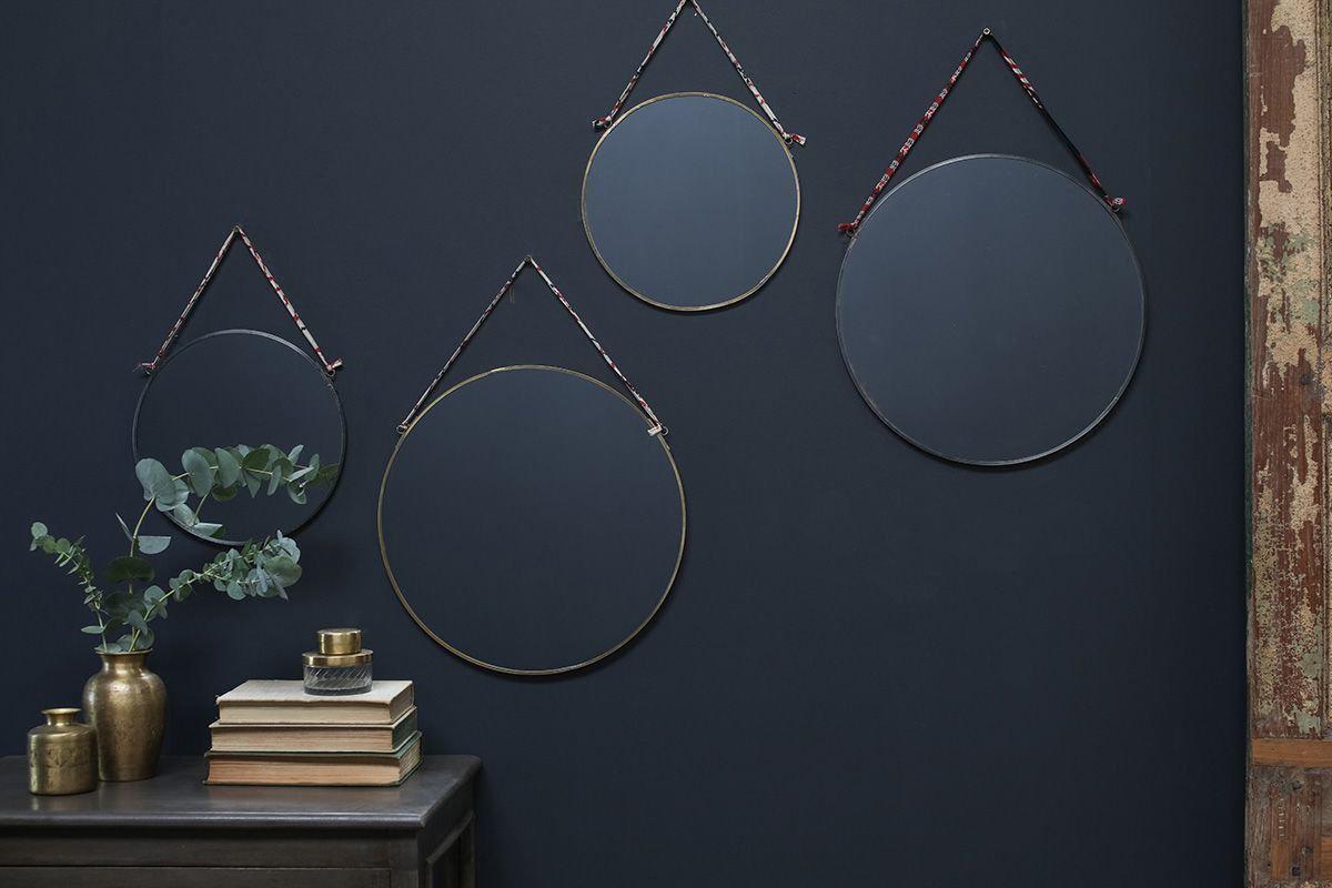 Four Kiko Round Mirror - Brass by nkuku are hung on a dark blue wall with recycled sari ties. Below, a dark table holds books, a plant in a gold vase, and gold accents. A weathered wooden panel appears at the right edge.