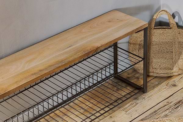 The Kiyoma Iron & Wood Low Standing Shelves by nkuku, featuring mango wood and a metal frame with two wire shelves, stands on a rustic wooden floor beside a straw tote bag, bringing an industrial touch to the space.
