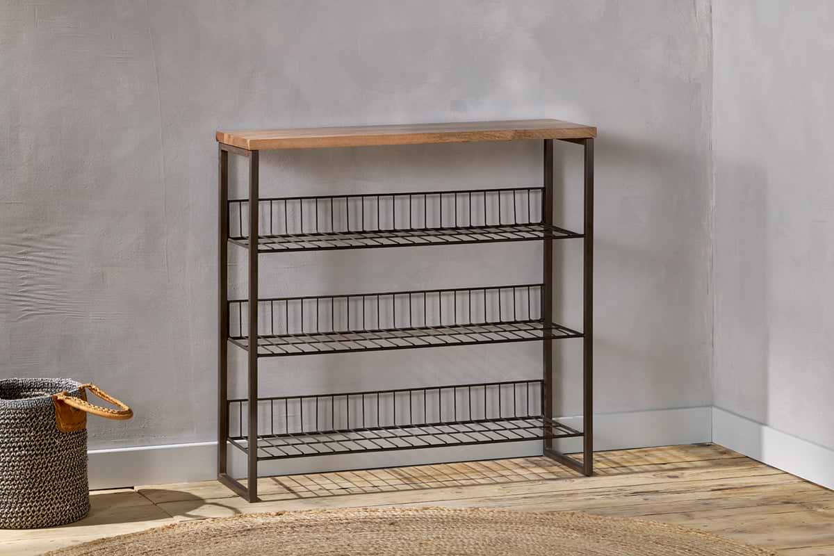 The nkuku Kiyoma Iron & Wood Tall Standing Shelves - Natural stand against a light gray wall on a wooden floor, combining minimalist and industrial style. A woven basket sits to the left, with a round woven rug partially in front.