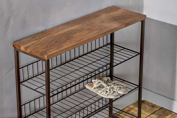 The Kiyoma Iron & Wood Tall Standing Shelves by nkuku, featuring a natural mango wood top and metal frame, stands against a light gray wall. Only a single pair of rope-strap sandals sits on the middle shelf; the others are empty on the wooden floor.