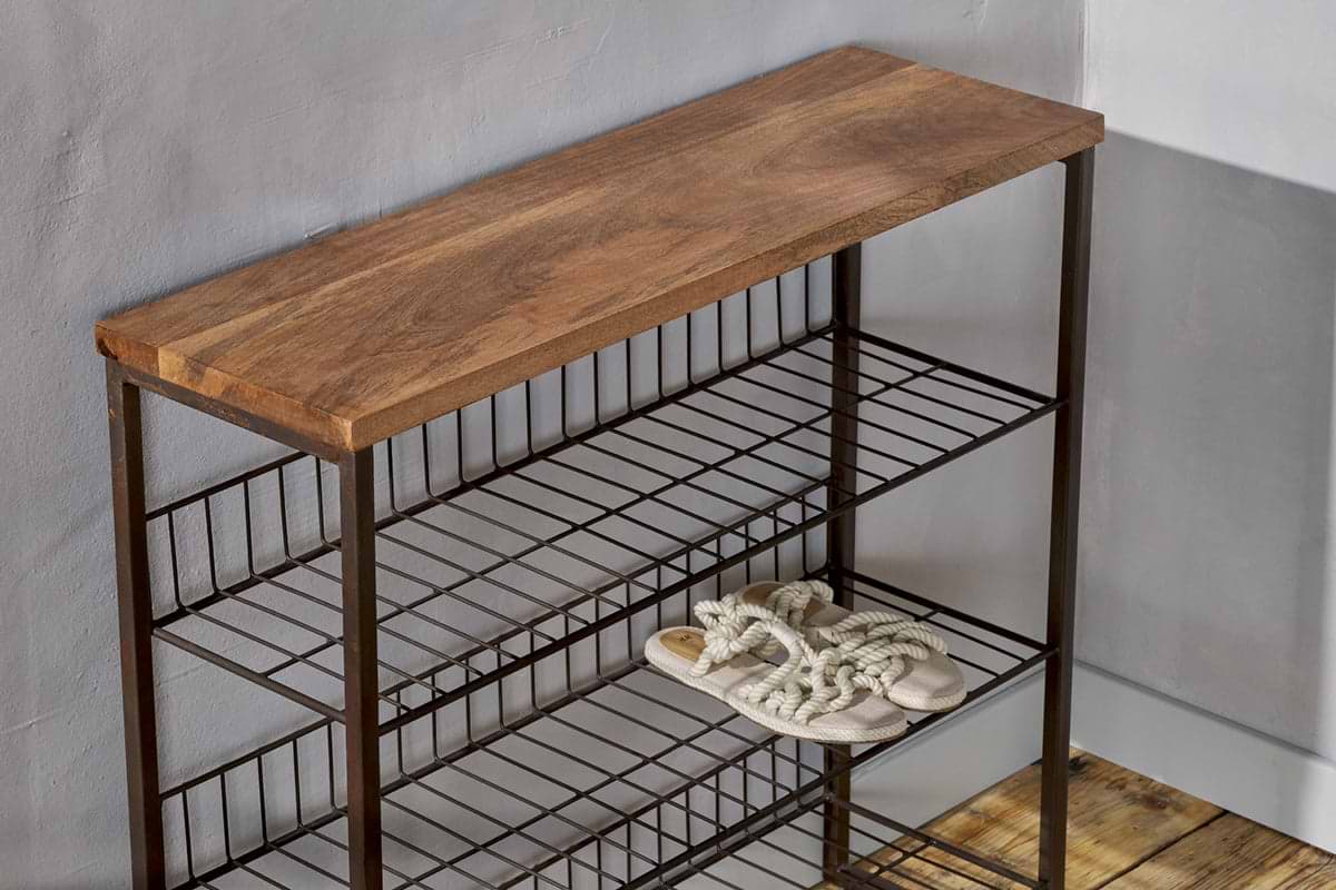 The Kiyoma Iron & Wood Tall Standing Shelves by nkuku, featuring a natural mango wood top and metal frame, stands against a light gray wall. Only a single pair of rope-strap sandals sits on the middle shelf; the others are empty on the wooden floor.