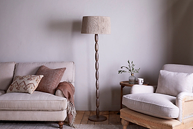 A cozy living room with a beige sofa and armchair, both adorned with neutral cushions and throws. Between them stands the nkuku Konali Mango Wood Floor Lamp in Ash Stain, featuring a textured shade that brings warmth in natural tones.