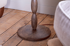 A close-up of the nkuku Konali Mango Wood Floor Lamp - Ash Stain highlights its circular base and rounded, turned stem on a wooden floor, with neutral upholstered furniture in the background for a cozy home lighting ambiance.