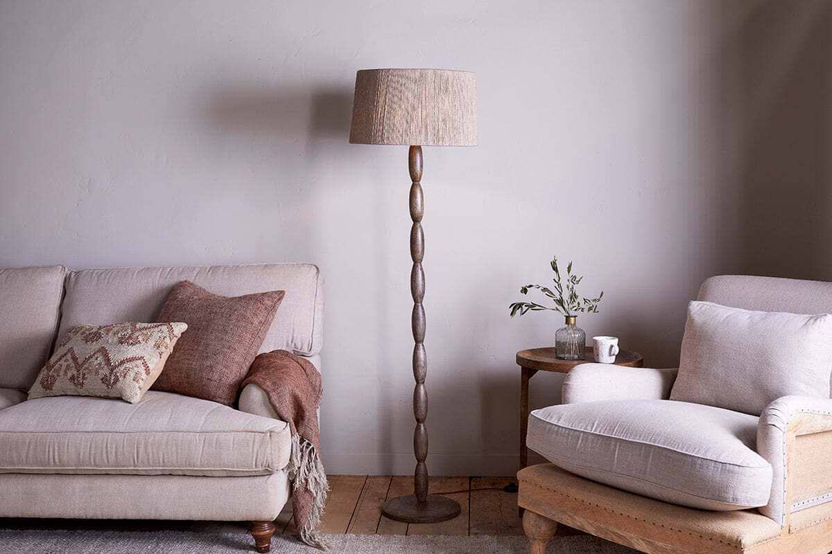 A cozy living room with a beige sofa, patterned pillow, and brown throw on the left, a tall nkuku Konali Mango Wood Floor Lamp - Ash Stain in the center, and a cushioned beige armchair with a small side table holding a plant and mug on the right.