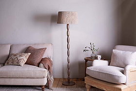 A cozy living room with a beige sofa, patterned pillow, and brown throw on the left, a tall nkuku Konali Mango Wood Floor Lamp - Ash Stain in the center, and a cushioned beige armchair with a small side table holding a plant and mug on the right.