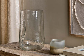 The Kotri Recycled Glass Organic Shape Vase - Clear by nkuku and a small frosted candle holder sit on a rustic wooden table, with a beige wall, framed artwork, and part of a closed book visible nearby.
