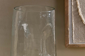 A close-up of the Kotri Recycled Glass Organic Shape Vase - Clear by nkuku set against a beige wall, with a partial view of framed textured fabric art to the right. The scene features a neutral, minimalist aesthetic.