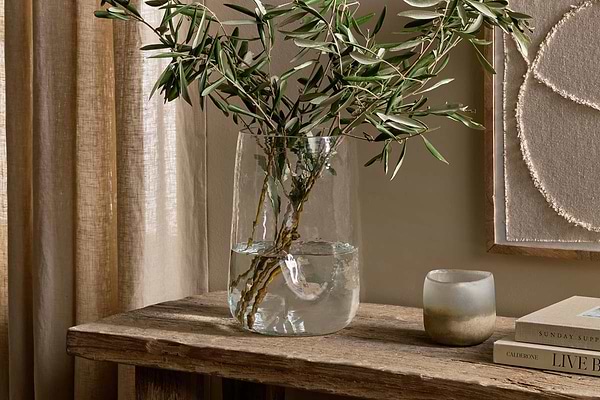 A Kotri Recycled Glass Organic Shape Vase - Clear by nkuku, filled with green branches and water, sits on a rustic table beside a frosted candle holder and stacked books, with beige curtains and textured wall art in the softly lit background.