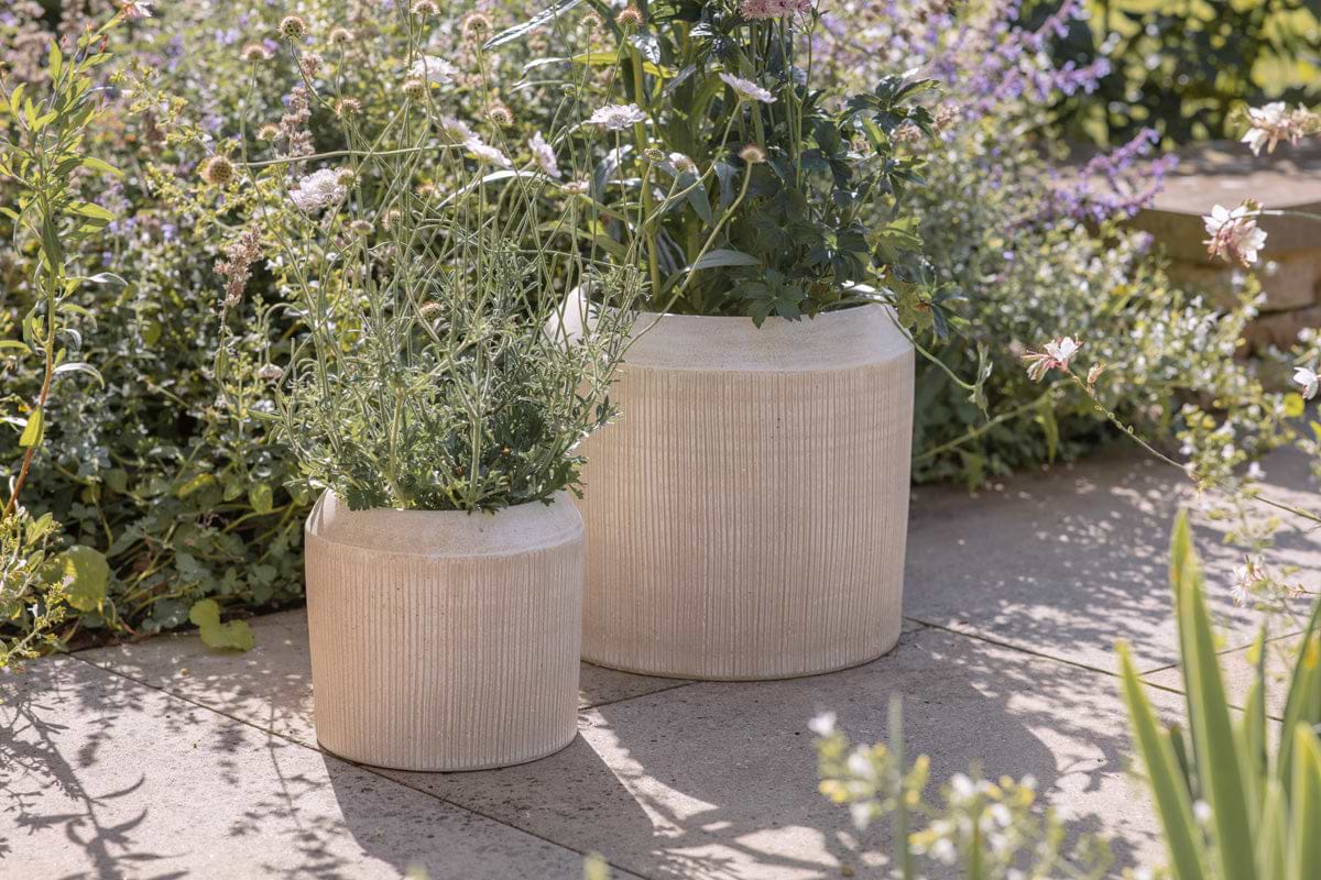 Two Kubo Ceramic Planters by nkuku in cream, featuring a textured reactive glaze, are displayed on a stone patio with green plants and pink flowers. Lush garden greenery surrounds these versatile indoor-outdoor planters.