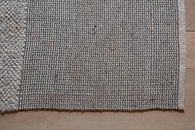 A close-up of the nkuku Kufri Jute Rug in Soft Blue & Brown shows its rectangular, textured woven surface and clearly defined edges. The handcrafted rug features a raised pattern along the left, resting on a light wood floor.