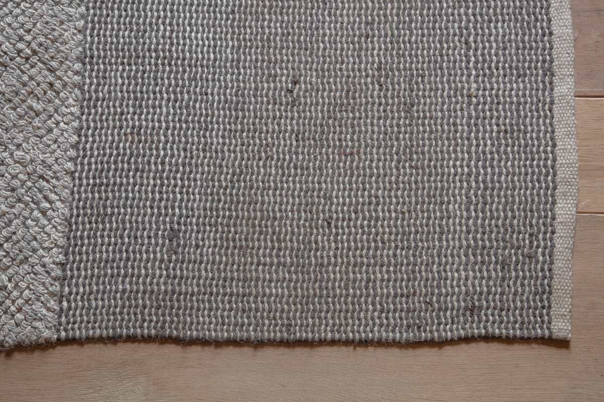 A close-up of the nkuku Kufri Jute Rug in Soft Blue & Brown shows its rectangular, textured woven surface and clearly defined edges. The handcrafted rug features a raised pattern along the left, resting on a light wood floor.