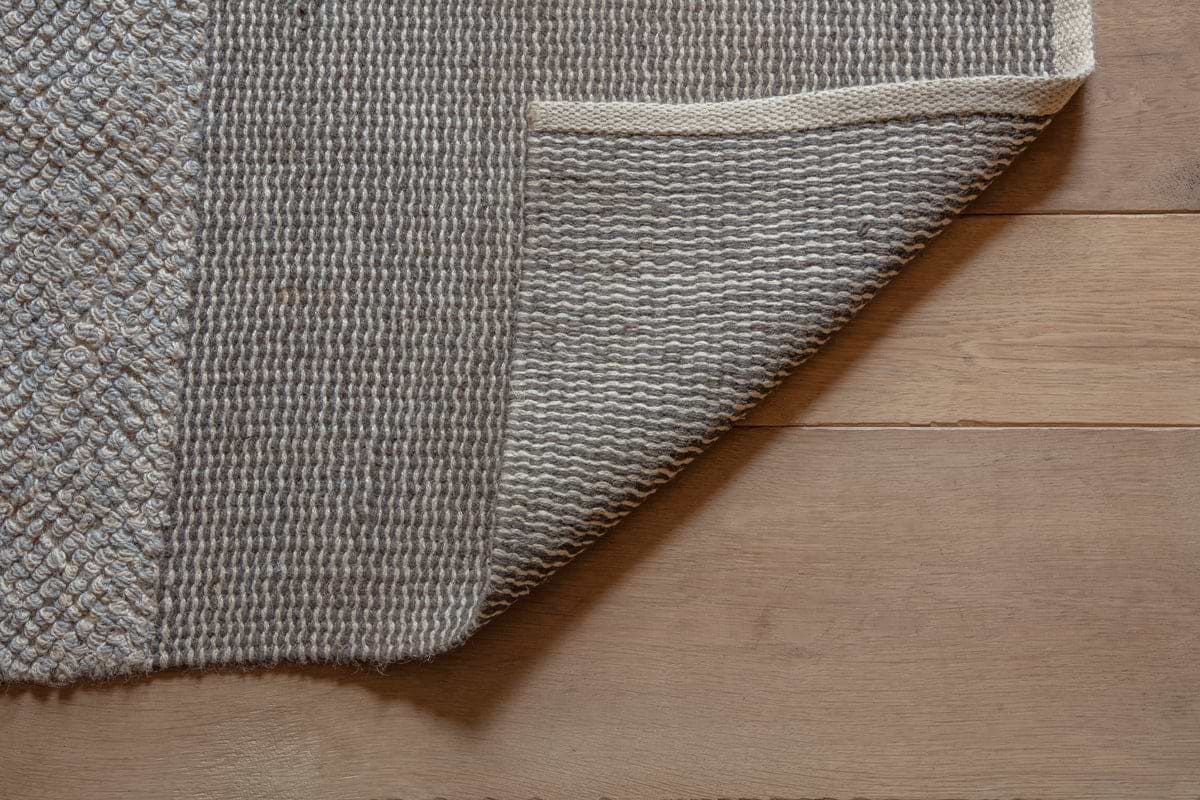 The Kufri Jute Rug by nkuku, in soft blue and brown, is partially flipped on a light wooden floor, showing both its textured top and underside. The floorboards run horizontally beneath the rug’s handcrafted design.
