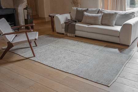 A cozy living room with a light gray sofa, matching pillows, and a brown throw. A woven armchair sits beside the nkuku Kufri Jute Rug in Soft Blue & Brown on wooden flooring, while sunlight filters through floor-length curtains.