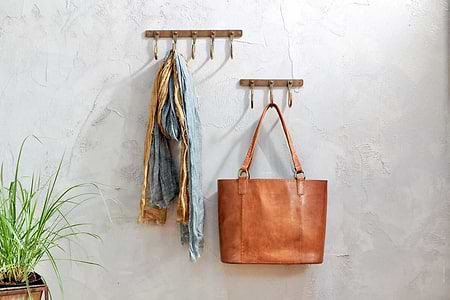 Two Kuna Iron Hooks in Antique Brass by nkuku adorn a textured gray wall. The left hook displays blue and gold scarves, while the right holds a tan leather tote—perfect for organizing small items in style.