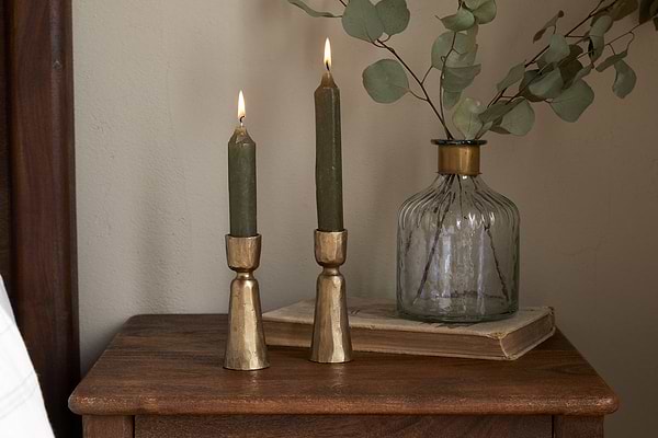 Two green candles in nkukus Lokoja Candle Holder - Antique Brass (Set of 2) sit on wood beside a ribbed glass vase with eucalyptus and a closed book, creating a cozy, rustic decor scene.