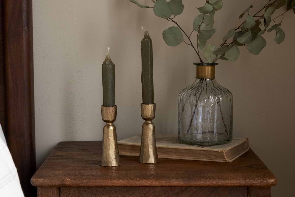Two green candles in nkukus Lokoja Candle Holder - Antique Brass (Set of 2) sit on a wooden table beside a clear glass vase with eucalyptus branches and a closed book, set against a neutral-toned wall.