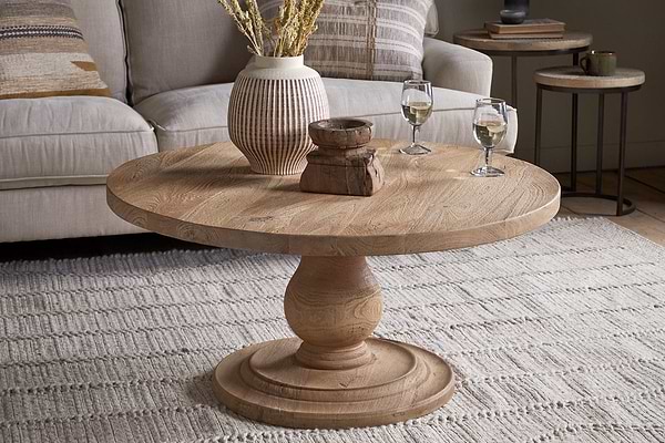The Laya Round Carved Coffee Table - Natural by nkuku, with its sculptural form and raw wood finish, sits on a textured rug. On top are a ceramic vase with dried flowers, two wooden decor pieces, and two glasses of white wine. A beige sofa is in the background.